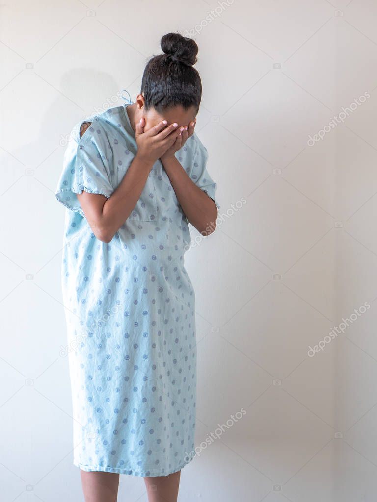 A young pregnant mixed race African American woman wearing a hospital gown or robe puts or hides her face in her open palms as she hunches over crying tears of sadness or depression.