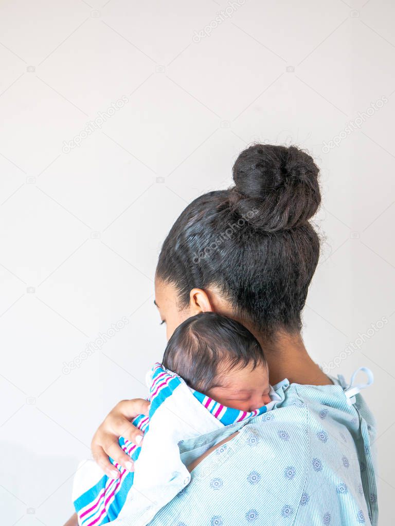 A mixed race African American mother faces away from the camera wearing a hospital gown holds her brand new infant baby over her shoulder hugging and cradling him as he sleeps in his blanket swaddle.