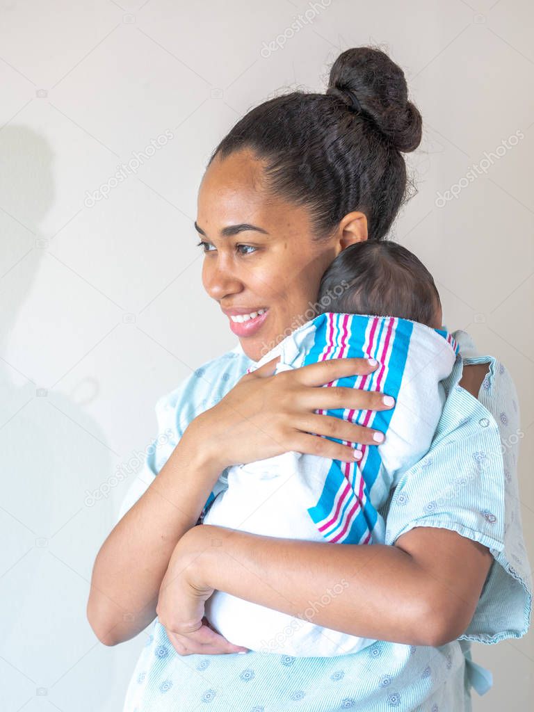 A happy mixed race African American mother wearing a hospital gown holds her new healthy infant baby boy on her shoulder hugging and cradling him as she looks to the side as he sleeps in his swaddle.