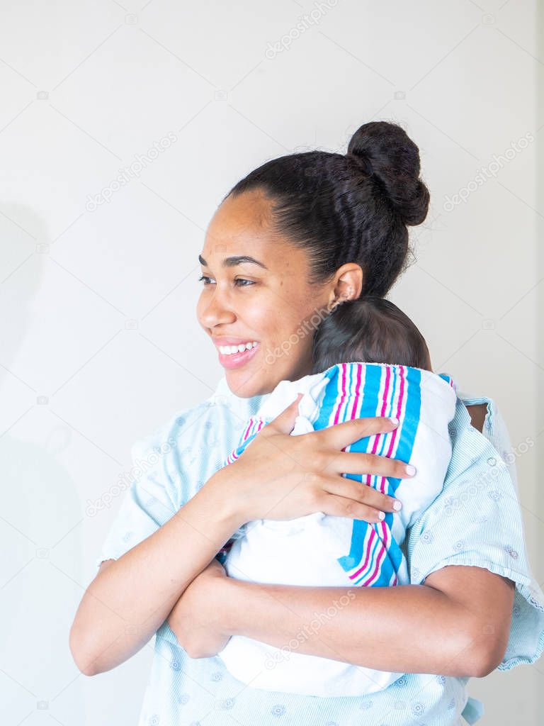 A happy mixed race African American mother wearing a hospital gown holds her new healthy infant baby boy on her shoulder hugging and cradling him as she looks to the side as he sleeps in his swaddle.