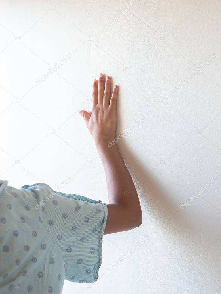 A closeup of a female medical patient wearing a hospital gown bracing her arm and open hand against the wall for support as she is in pain or agony.