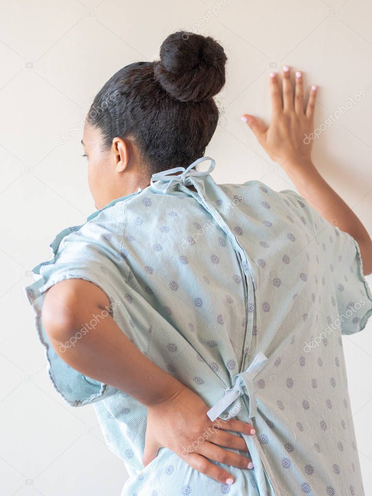 A close up photograph of a middle aged African American mixed race woman with her hand on her lower back or hip hunched over in pain and other hand on the wall wearing a patterned hospital gown.