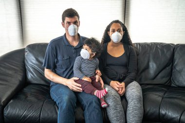 Genç ve melez bir aile, karonavirüs ya da Covid-19 hastalığından korunmak umuduyla oğullarını siyah deri bir kanepeye oturtup yüzlerine toz maskesi takıyor..
