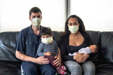 Karışık bir aile siyah deri bir koltukta oturur. Anne ve baba, anne ve oğullarını kucaklar. Hepsi de karonavirüs veya Covid-19 hastalığından korunmak umuduyla tıbbi maske takar.