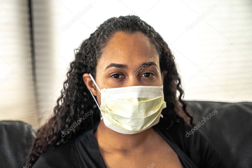 A close up portrait of a beautiful young African American mother sitting on a sofa in her home wearing a medical face mask in hopes of preventing getting sick from caronavirus or COVID-19.