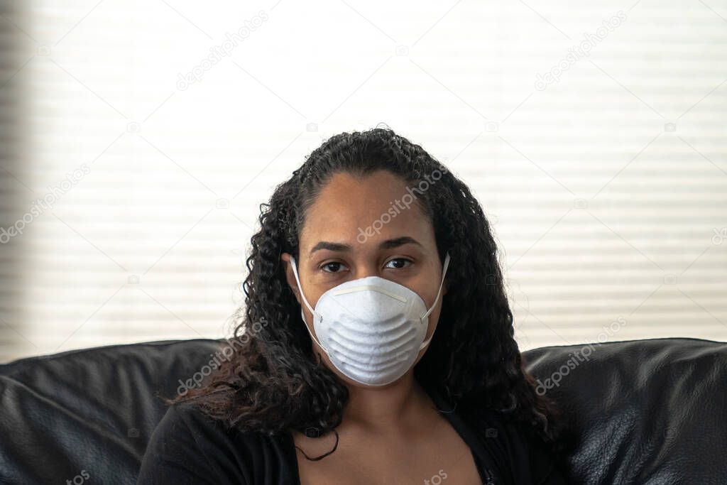 A close up portrait of a beautiful young African American mother sitting on a sofa in her home wearing a dust face mask in hopes of preventing getting sick from caronavirus or COVID-19.