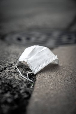 Kirli bir tıbbi maske ya da cerrahi solunum maskesi yakın plan atılmış ve çöp olarak asfalt sokakta beton kaldırım ve kanalizasyon kanalizasyonunun yanına bırakılmış..