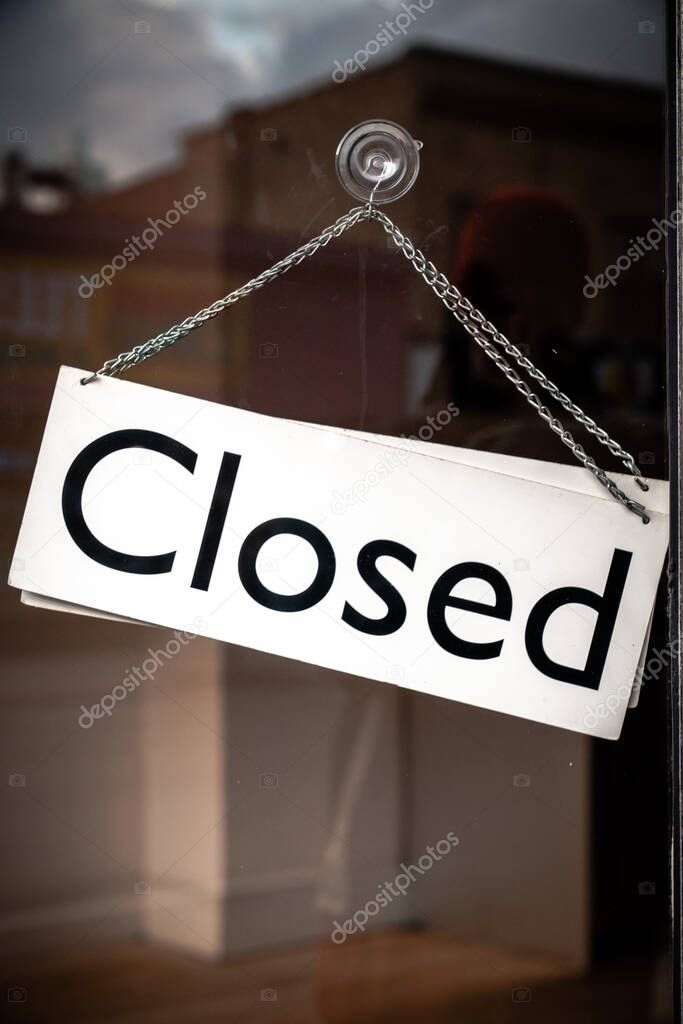 Classic black and white sign and chain hangs on an angle in a glass storefront or door due to the shop or retail business being closed during the COVID-19 outbreak and government stay at home orders.