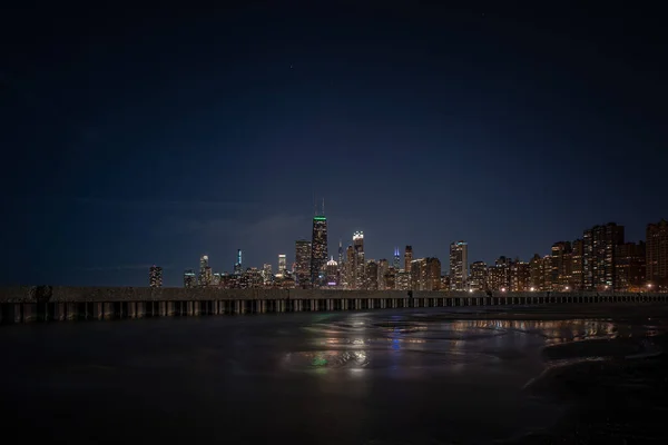 Building lights reflecting off of standing water adjacent to corrugated steel sheet piling and concrete pier with the Chicago Skyline on the horizon and stars and planes in the sky above.