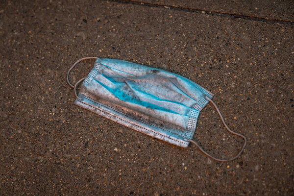 A close up of a lost, discarded or dropped blue single worn, dirty and wet face mask or surgical doctor or nurse mask with elastic bands littering the sidewalk or concrete pavement in downtown Chicago