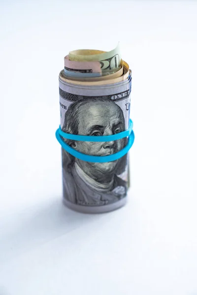 Close up photograph of a rolled up wad of cash sitting on edge with a blue rubber band wrapping across Benjamin Franklin\'s face on a hundred dollar bill on a white background with copy space.