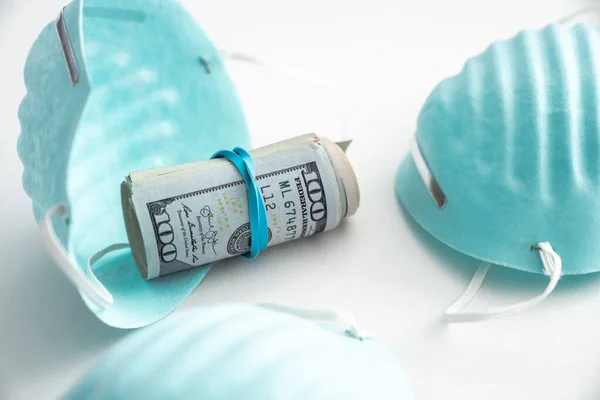 A rolled up wad of cash with a rubberband around it sits in a blue ribbed medical mask or protective face covering used for COVID-19 virus pandemic isolated on a white background.