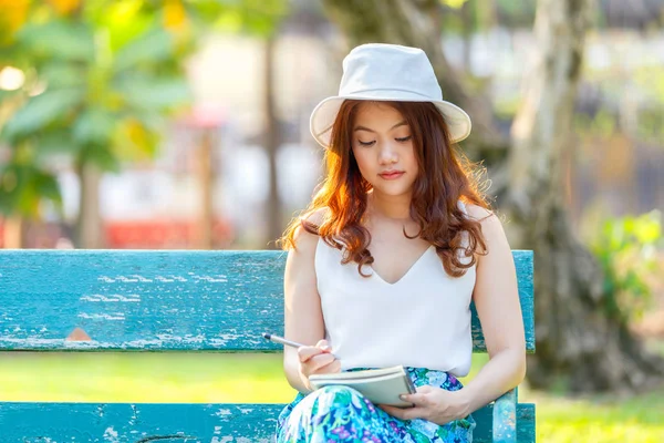Mulheres asiáticas bonitas com caneta escrevendo caderno e sentado em um banco de madeira e em um parque — Fotografia de Stock