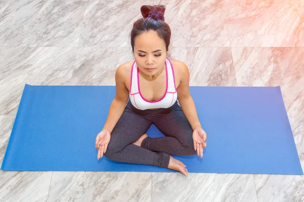 Aantrekkelijke jonge vrouw thuis uit te werken, doen yoga oefening op blauwe mat, zittend in gemakkelijk fatsoenlijk. — Stockfoto