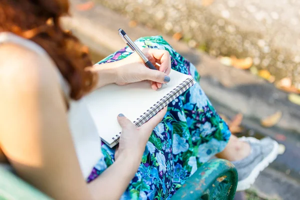 Close up left hand asian women with pen writing notebook and sitting on a wooden bench and a park. — Stock Photo, Image