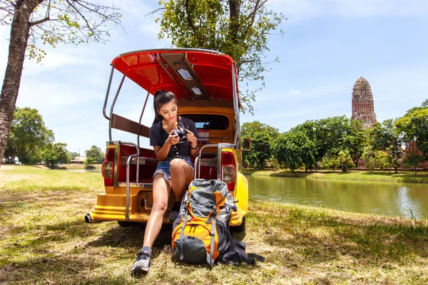 Young asian female traveler with backpack traveling sitting on taxi or Tuk Tuk and seeing photo on vintage camera with Wat Mahathat background, Ayutthaya Province, Thailand