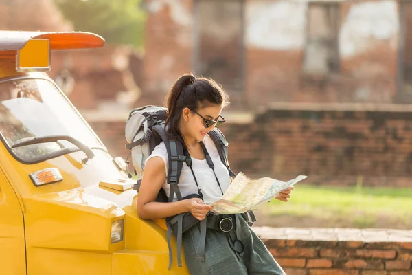 Young asian female traveler with backpack traveling standing on taxi or Tuk Tuk and see map travel with old temple background, Ayutthaya Province, Thailand