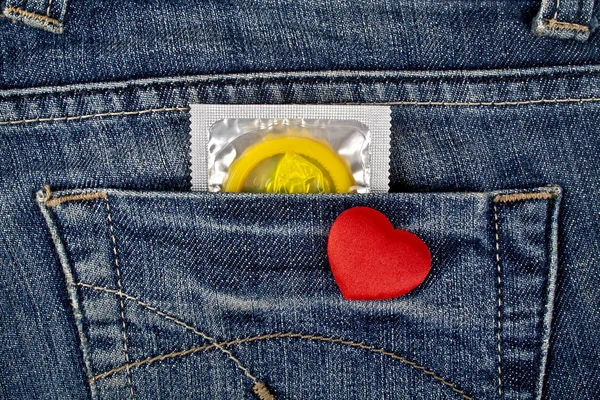 Red heart and yellow condom in blue jeans pocket — Stock Photo, Image