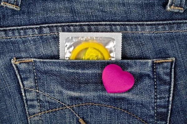 Violet heart and yellow condom in blue jeans pocket — Stock Photo, Image