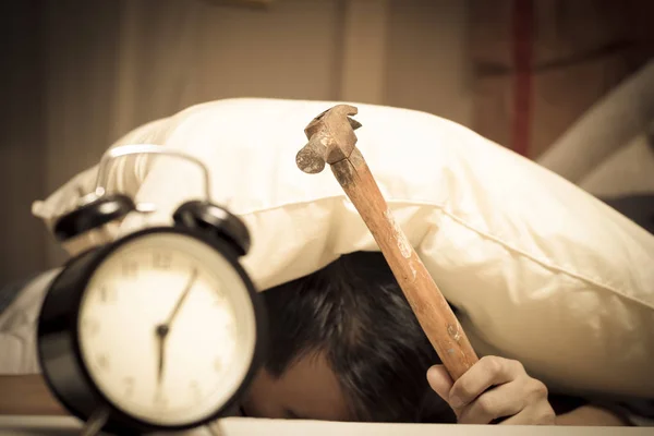 Schlafender asiatischer junger Mann wird frühmorgens durch Wecker auf Bett gestört & hält Hammer in der Hand — Stockfoto