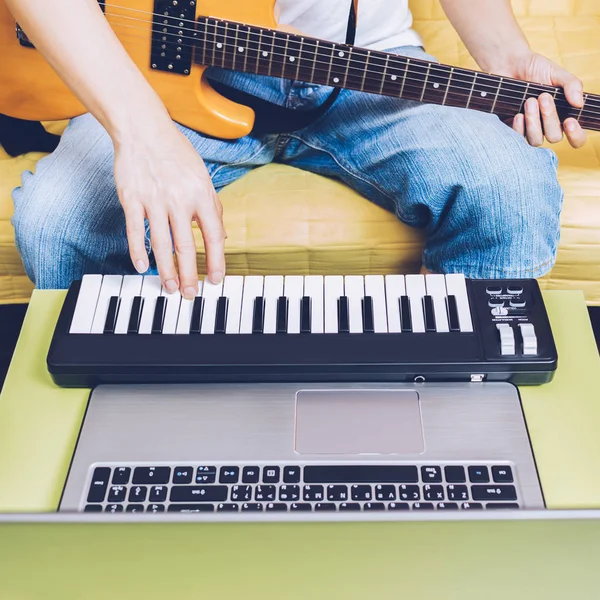 Musicien masculin composant une chanson sur ordinateur portable, clavier midi et guitare électrique dans le salon pour la technologie de production de musique à la mode dans le mode de vie moderne Image En Vente