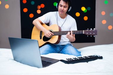 online müzik dersi beyaz yatakta gitarla geceleri pratik dizüstü bilgisayar ve MIDI klavye ile bir şarkı bestelemek Asya erkek müzisyen. Şarkı yazma, müzik eğitimi ve yatak odası stüdyo konsepti