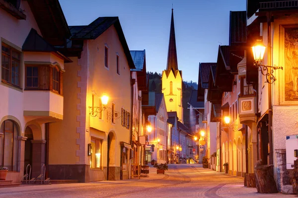 Empty main street in blue hour in historic center of Bavarian resort town Garmisch-Partenkirchen with spire of Maria Himmelfahrt catholic church above row of traditional German houses, Bayern Germany Europe