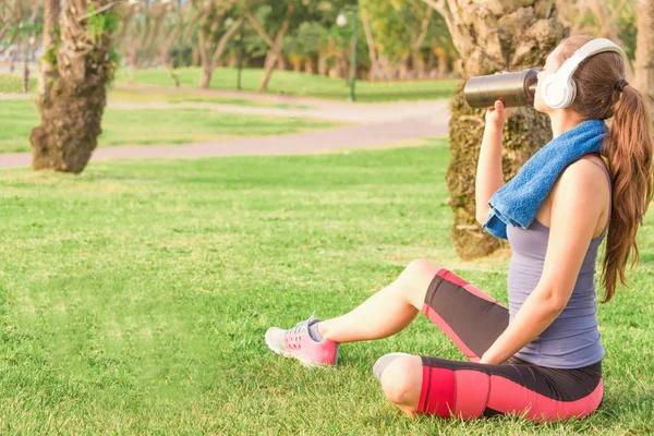 Una giovane ragazza in un parco sull'erba Dopo un allenamento di fitness con le cuffie e beve frullato proteico Foto Stock
