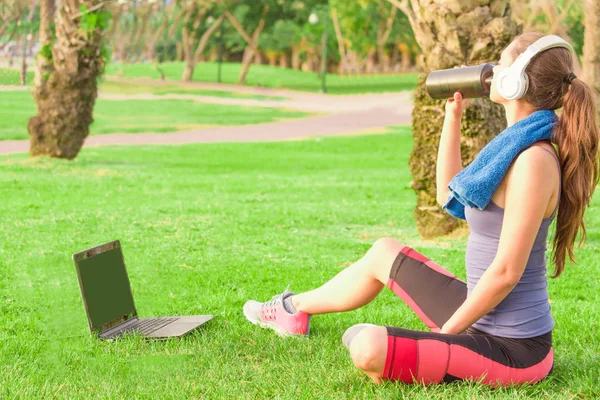 Una giovane ragazza in un parco sull'erba Dopo un allenamento di fitness con laptop e cuffie e beve frullato proteico Immagine Stock