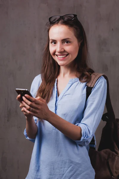 Retrato de mujer sonriente con gafas y teléfono celular cerca de la pared de fondo gris —  Fotos de Stock