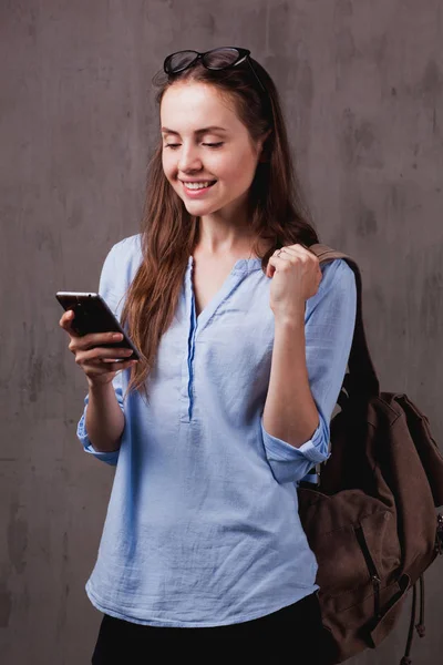 Retrato de mujer sonriente con gafas y teléfono celular cerca de la pared de fondo gris —  Fotos de Stock