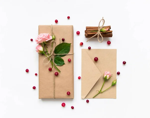 Gift box and envelope in eco paper on white background. Presents decorated with roses and berries. Holiday concept, top view, flat lay