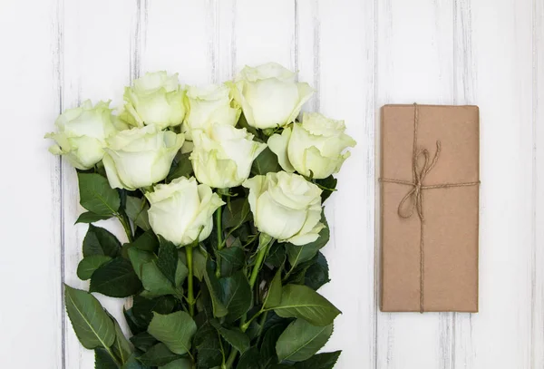 Strauß luxuriöser weißer Rosen und eine Geschenkschachtel aus Ökopapier auf weißem Holzhintergrund. Draufsicht, flache Lage — Stockfoto