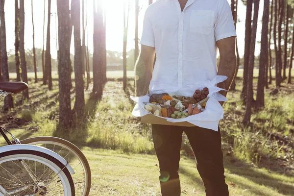 Guy holding a mixed food platter with fruit, meat and cheese in a park at sunset