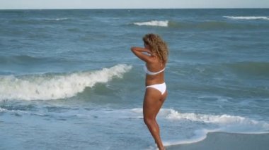 Genç güzel seksi tabaklanmış kız bikinili kumsalda gün batımında deniz kenarında oturan kıvırcık saçlı. O obscurities gülümsedi, kendini ve yavaş hareket okşadı. 120fps