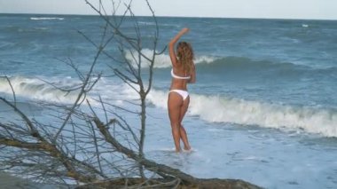 Genç güzel seksi tabaklanmış kız bikinili kumsalda gün batımında deniz kenarında oturan kıvırcık saçlı. O obscurities gülümsedi, kendini ve yavaş hareket okşadı. 120fps
