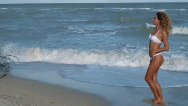 Joven hermosa chica bronceada sexy en bikini con el pelo rizado sentado en la playa al atardecer junto al mar. Sonrió enigmáticamente, se acarició y... En cámara lenta. 120 fps — Vídeo de stock