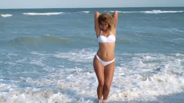 Jonge mooie sexy gelooid meisje in bikini met krullend haar zittend op het strand bij zonsondergang aan zee. Ze glimlachte raadselachtig, streelde zichzelf en Slow-motion. 120fps — Stockvideo