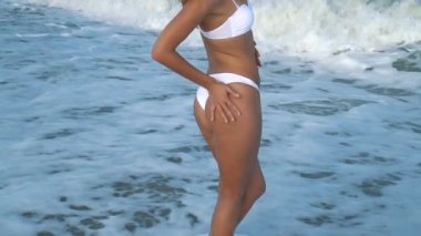 Genç güzel seksi tabaklanmış kız bikinili kumsalda gün batımında deniz kenarında oturan kıvırcık saçlı. O obscurities gülümsedi, kendini ve yavaş hareket okşadı. 120fps