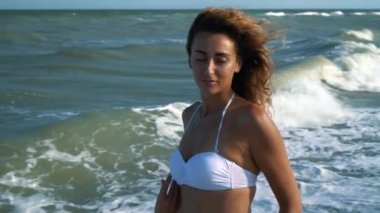 Genç güzel seksi tabaklanmış kız bikinili kumsalda gün batımında deniz kenarında oturan kıvırcık saçlı. O obscurities gülümsedi, kendini ve yavaş hareket okşadı. 120fps