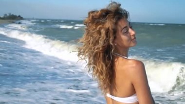 Genç güzel seksi tabaklanmış kız bikinili kumsalda gün batımında deniz kenarında oturan kıvırcık saçlı. O obscurities gülümsedi, kendini ve yavaş hareket okşadı. 120fps