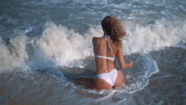 Joven hermosa chica bronceada sexy en bikini con el pelo rizado sentado en la playa al atardecer junto al mar. Sonrió enigmáticamente, se acarició y... En cámara lenta. 120 fps — Vídeos de Stock