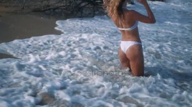 Genç güzel seksi tabaklanmış kız bikinili kumsalda gün batımında deniz kenarında oturan kıvırcık saçlı. O obscurities gülümsedi, kendini ve yavaş hareket okşadı. 120fps