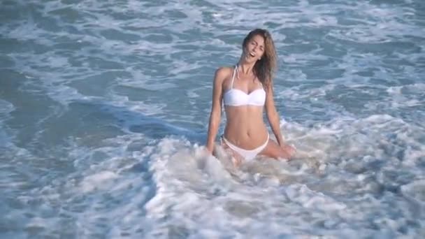 Krásná mladá sexy opálená dívka v bikinách s kudrnatými vlasy sedí na pláži při západu slunce u moře. Záhadně usmál, pohladil sama a. zpomaleně. 120fps — Stock video
