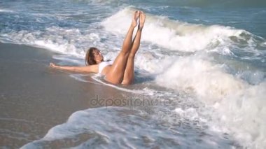 Genç güzel seksi tabaklanmış kız bikinili kumsalda gün batımında deniz kenarında oturan kıvırcık saçlı. O obscurities gülümsedi, kendini ve yavaş hareket okşadı. 120fps