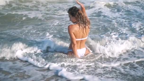 Menina bronzeada sexy bonita nova em biquíni com cabelo encaracolado sentado na praia ao pôr do sol junto ao mar. Ela sorriu enigmaticamente, acariciou-se e. Movimento lento. 120fps — Vídeo de Stock