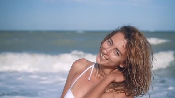 Jonge mooie sexy gelooid meisje in bikini met krullend haar zittend op het strand bij zonsondergang aan zee. Ze glimlachte raadselachtig, streelde zichzelf en Slow-motion. 120fps — Stockvideo