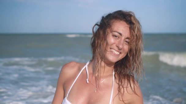 Menina bronzeada sexy bonita nova em biquíni com cabelo encaracolado sentado na praia ao pôr do sol junto ao mar. Ela sorriu enigmaticamente, acariciou-se e. Movimento lento. 120fps — Vídeo de Stock