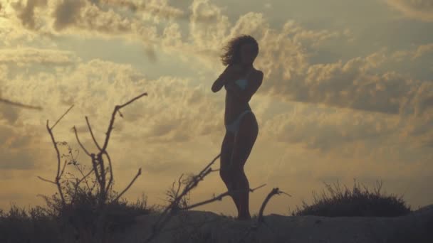 Młody piękny sexy Opalona dziewczyna w bikini z kręconymi włosami taniec na plaży o zachodzie słońca nad morzem. Ona uśmiechnął się enigmatycznie, gładził siebie i. zwolnionym tempie. 120 kl/s — Wideo stockowe