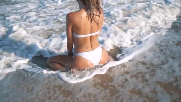 Joven hermosa chica bronceada sexy en bikini con el pelo rizado sentado en la playa al atardecer junto al mar. Sonrió enigmáticamente, se acarició y... En cámara lenta. 120 fps — Vídeos de Stock